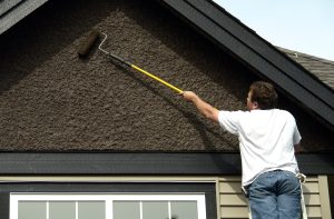 Man on ladder repainting stucco on house with long paint roller brush